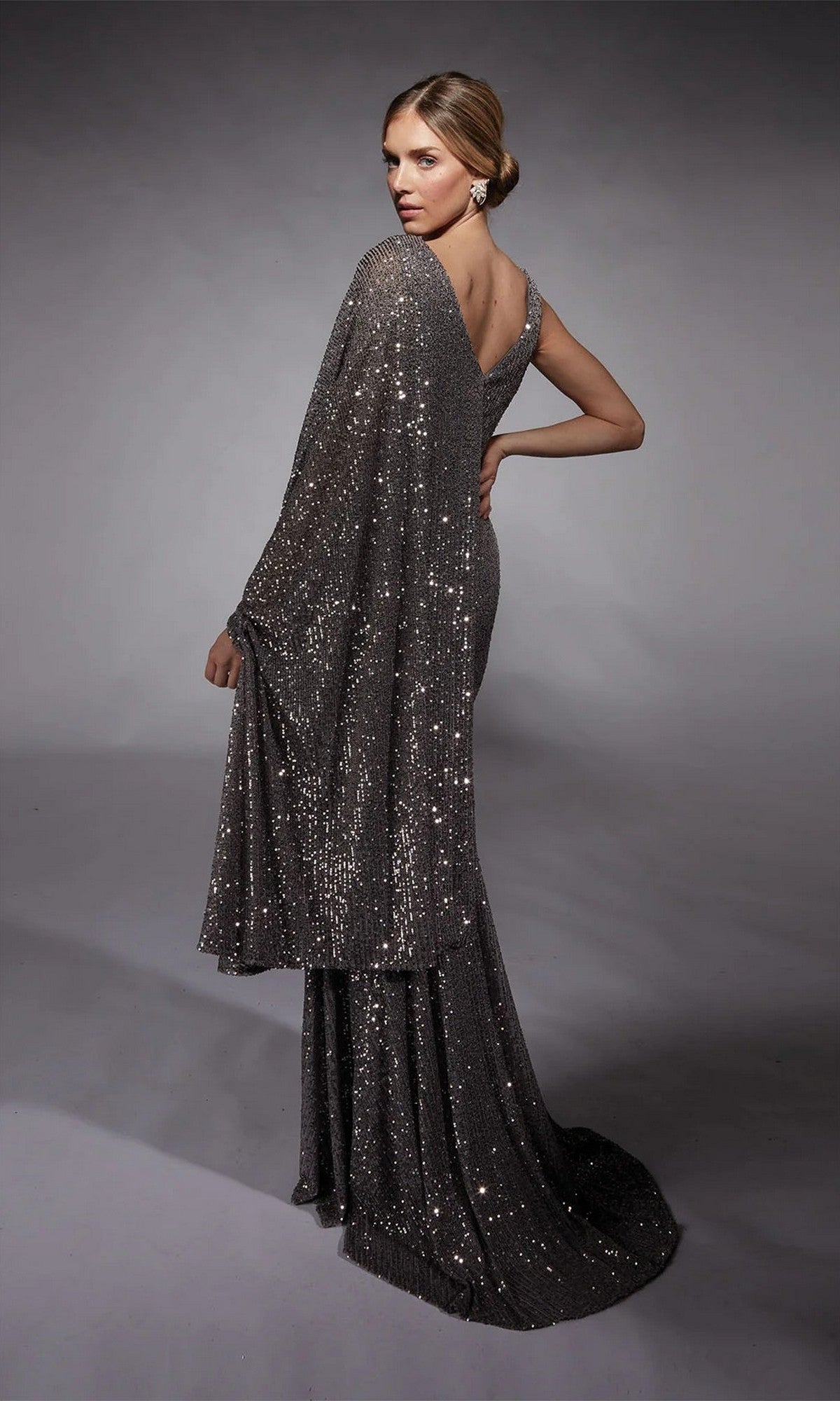 A woman poses elegantly against a neutral gray background wearing the Alyce 27718, a long formal dress featuring shimmering silver sequins and a matching shawl.